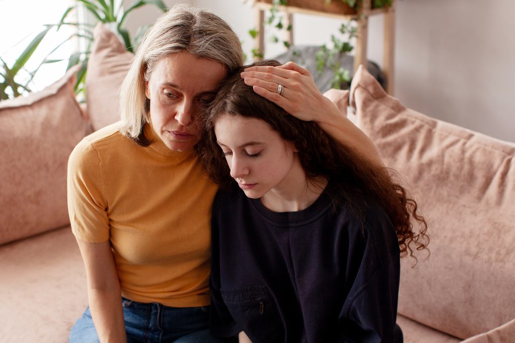 A mother sitting on a sofa talking calmly with her upset teenage daughter about teen substance use treatment.