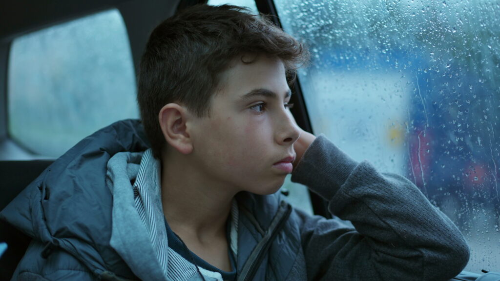 A teenage boy looking out a window with a flat affect, illustrating parent concerns about teens and psychiatric medications causing a loss of personality or feeling like a zombie.