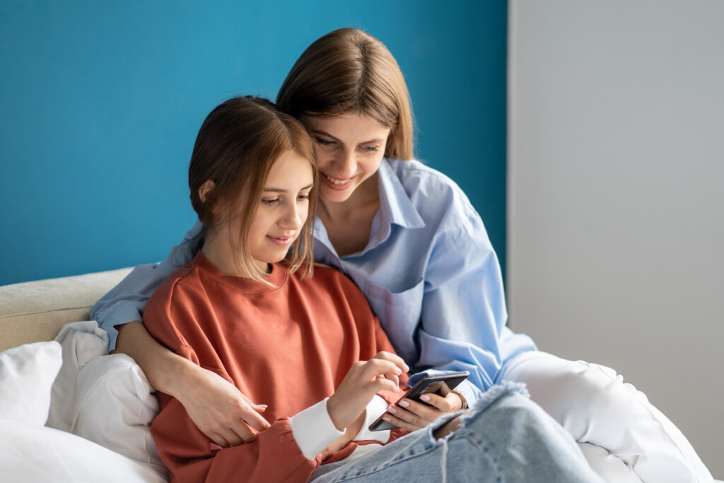 Permissive mom sitting with daughter watching videos on smartphone instead of going to school.