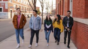 A group of young people engaged in lively conversation, walking down a city street lined with barren trees and red brick buildings.