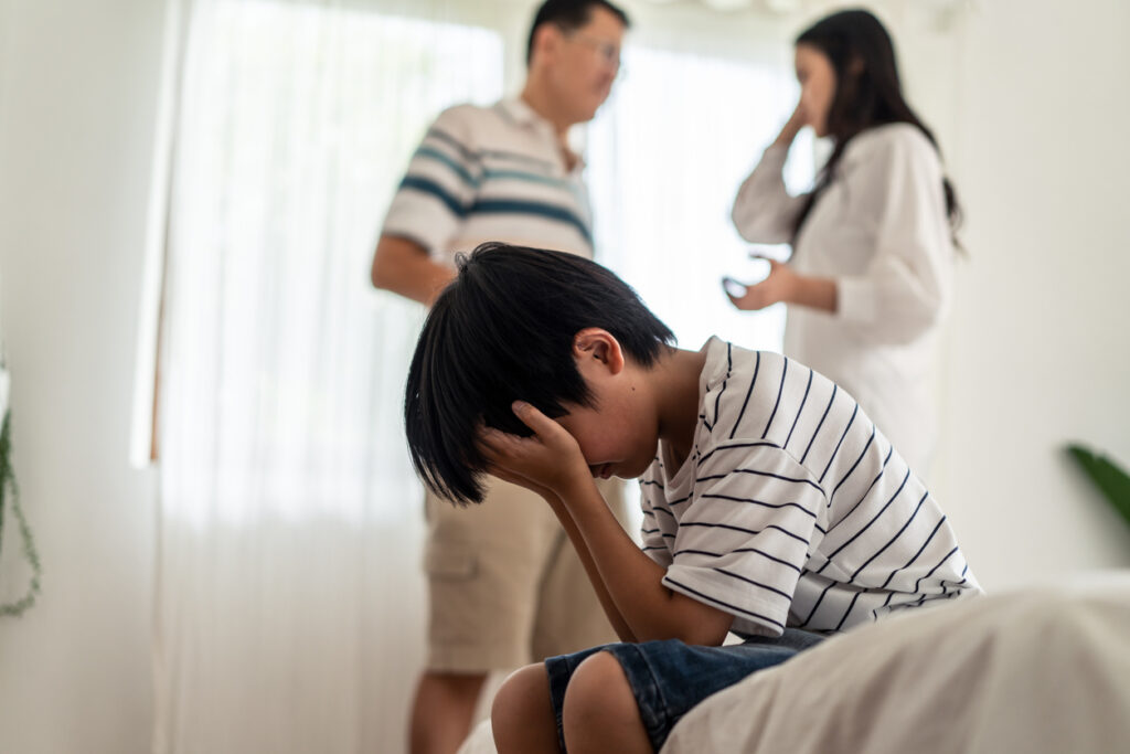 An upset teen holds his head into his hands and his parents are discussing getting a divorce