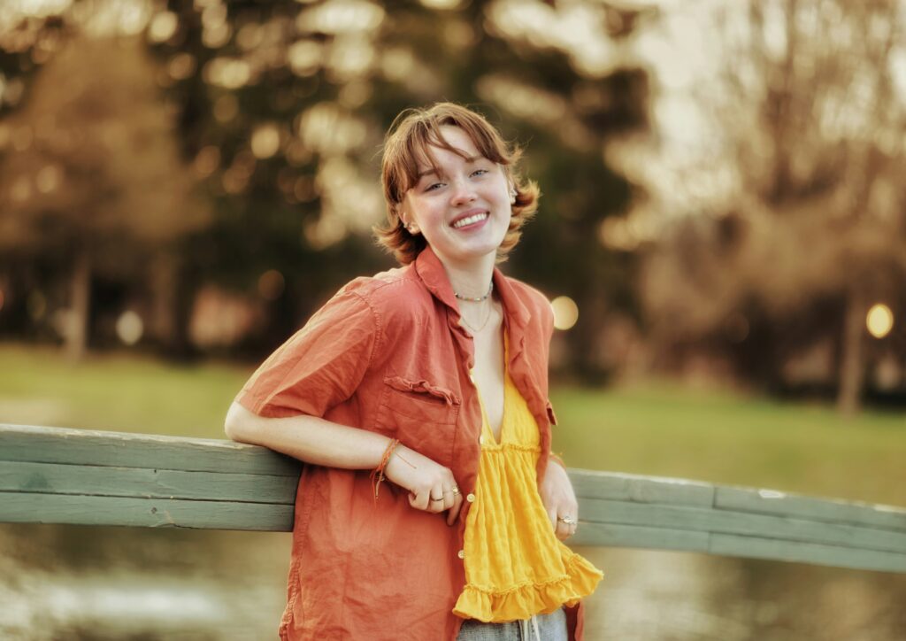 A young person with short, coppery hair and light skin wears an orange short-sleeve button-up, opened to reveal a bright yellow ruffle tank underneath. They lean against a wooden fence, smiling and gazing into the camera. The blurred background has yellowed trees and warm, natural light.