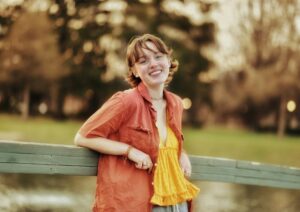 A young person with short, coppery hair and light skin wears an orange short-sleeve button-up, opened to reveal a bright yellow ruffle tank underneath. They lean against a wooden fence, smiling and gazing into the camera. The blurred background has yellowed trees and warm, natural light.