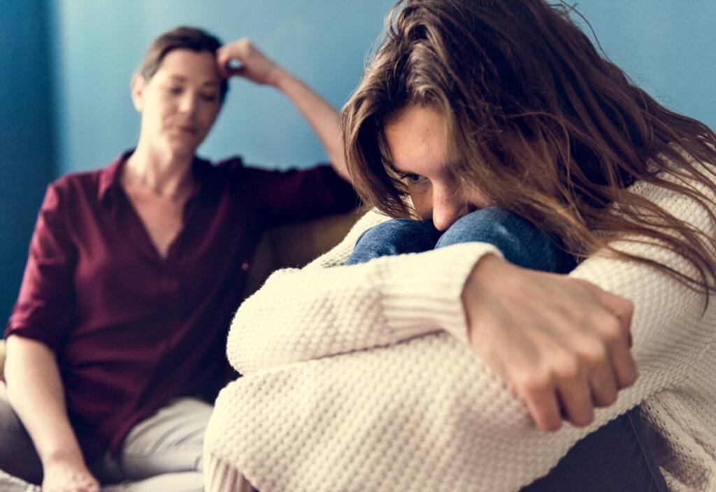 an upset teen hides her face as her mom looks concerned in the background